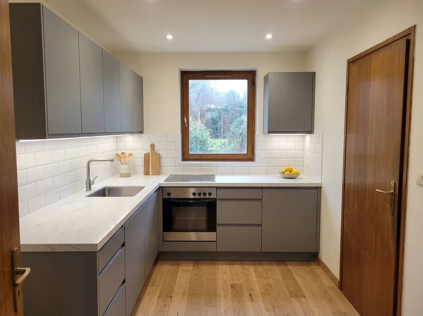 La même cuisine après travaux : façades gris mat sans poignée, plan de travail en quartz blanc, crédence métro et parquet chêne clair ; fenêtre et porte inchangées.